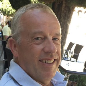image002 A middle-aged man smiles while sitting at an outdoor caffe table with a glass of white wine in the background.