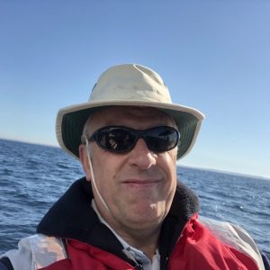 image003 A man wearing a hat, sunglasses, and a red jacket takes a selfie on a boat with the ocean and clear sky in the background.